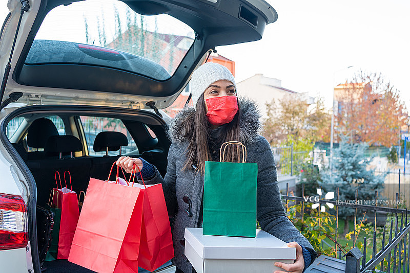 年轻美丽的微笑女子戴着预防疾病的防护面罩，从汽车后备箱卸下购物袋在她的房子停车场在圣诞节图片素材