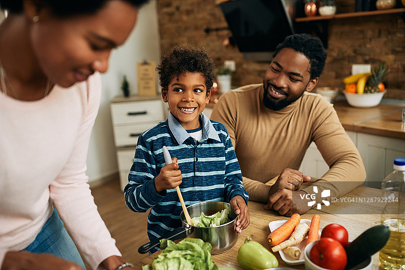 快乐的黑人男孩和他的父母在厨房准备健康的食物。图片素材