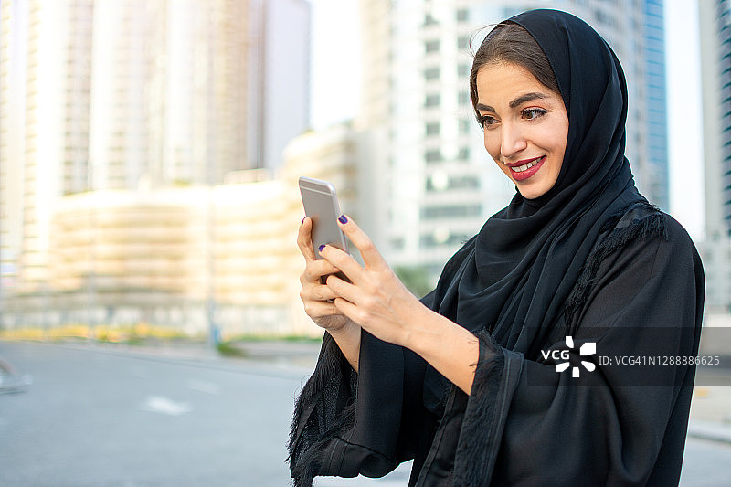 美丽微笑的年轻阿拉伯妇女穿着传统阿拉伯长袍在城市街道上使用智能手机的肖像。图片素材