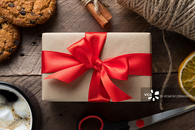 准备一个家庭假日的概念，快乐的平安夜。用鲜红的丝带打包并绑好圣诞礼物，还有带蝴蝶结的盒子，放在木桌的背景上。一杯热咖啡，棉花糖，饼干和巧克力。图片素材