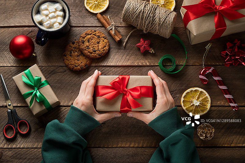 一个穿着绿色毛衣的女孩或女人拿着一份圣诞礼物。一家人准备一个概念节日，平安夜快乐。杯子里有热咖啡和棉花糖，木桌背景上有巧克力饼干。图片素材