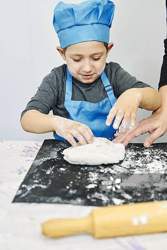 男孩扮演厨师，戴着厨师帽，围着蓝围裙做披萨面团图片素材