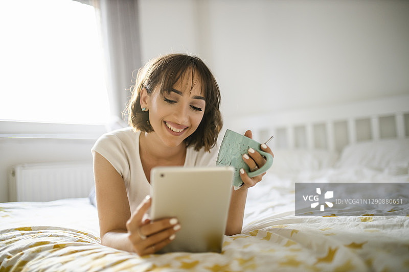 一个漂亮的年轻女子正在喝一杯热饮，并在床上使用她的数字平板电脑图片素材