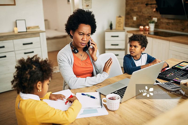 非裔美国母亲和两个孩子在家里一边用笔记本电脑工作一边打电话。图片素材