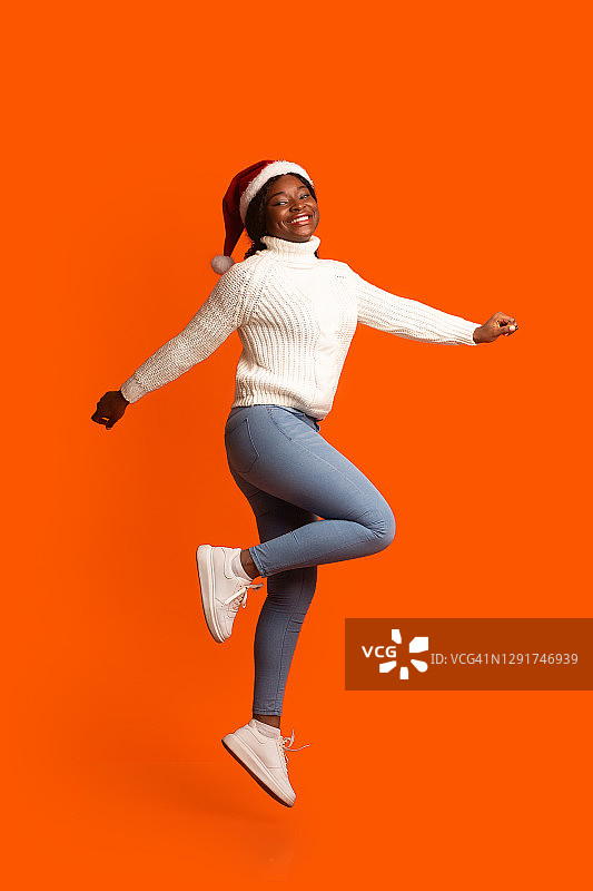 女性的圣诞老人。快乐的黑人妇女在圣诞节的帽子跳过橙色背景图片素材