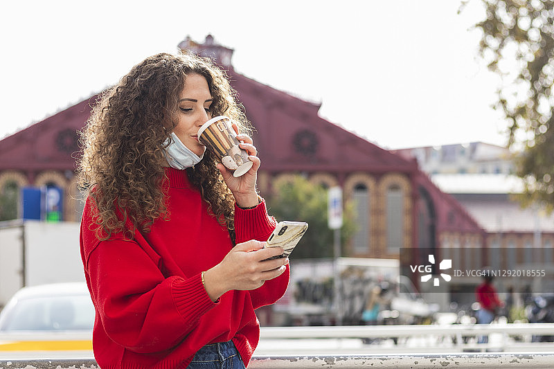 2019冠状病毒病期间，年轻女性在城市使用手机时喝咖啡图片素材
