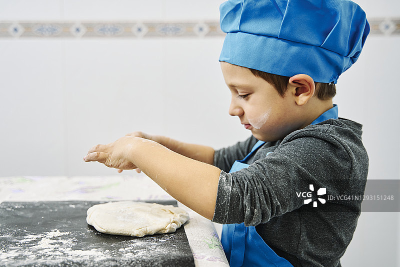 小男孩在厨房里玩厨师的游戏，戴着蓝色的帽子和围裙，做一个披萨面团图片素材