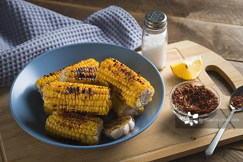 在一张木桌的背景下，一个明亮的陶瓷盘子上放着烤好的黄色玉米棒，上面撒着香料。自制食物的概念。图片素材
