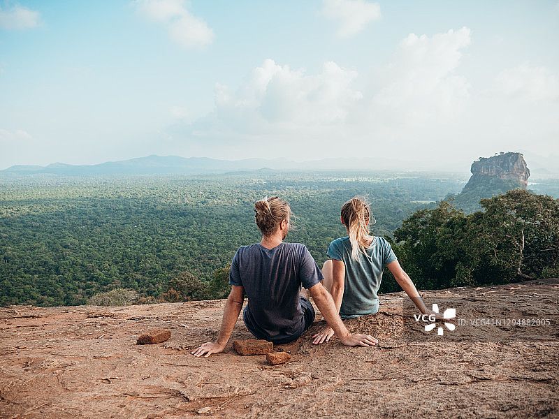 后视图的年轻夫妇坐在顶部的岩石在日落凝视美丽的丛林自然景观图片素材