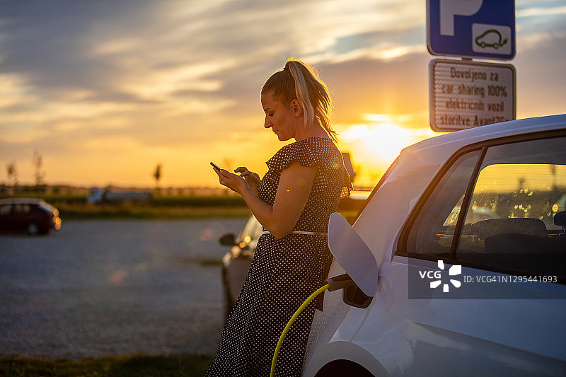 侧视图腰部以上的照片，一个金发成熟的女人与马尾使用她的手机，而等待她的白色电动车充电在日落图片素材