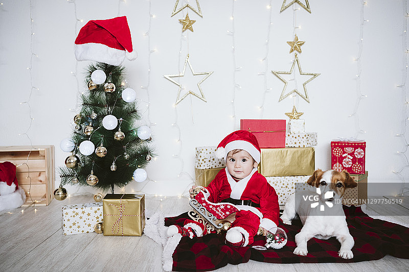 在圣诞节期间，一个穿着圣诞老人服装的小男孩坐在家里的狗旁边玩耍图片素材