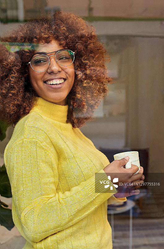 微笑的女人拿着咖啡杯，在咖啡馆的玻璃后面做着白日梦图片素材