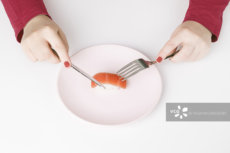 从上面，一个粉红指甲，深粉色毛衣，面目人非的女人正准备用粉色盘子里的餐具吃一片鲑鱼寿司。孤独的概念图片素材