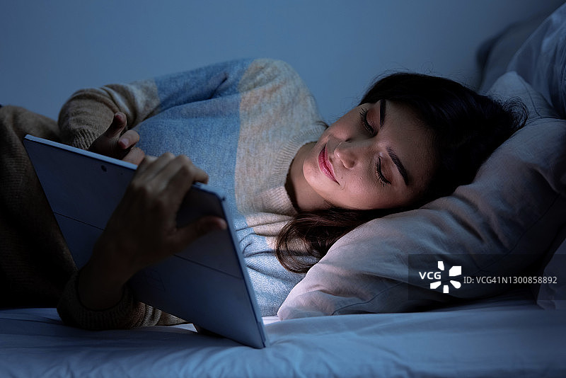 白种女性晚上睡觉前在床上使用平板电脑，手机成瘾概念，蓝光对眼睛有害。图片素材