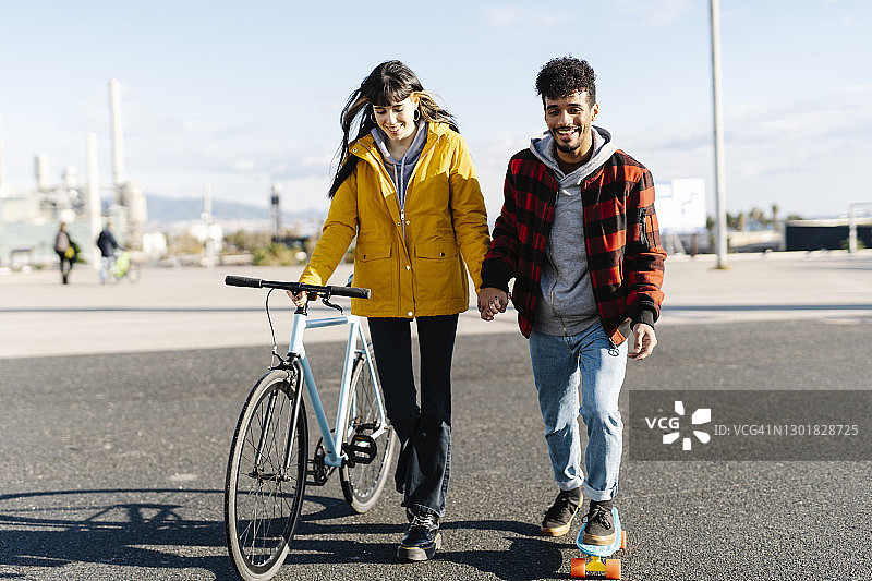 微笑的男子滑板，而牵着朋友的手与自行车走在路上图片素材