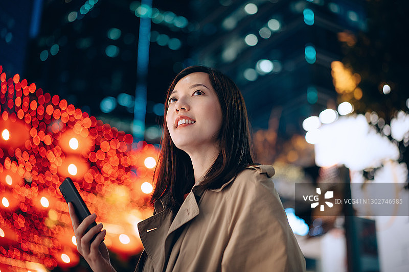 这是一幅低角度的亚洲女商人在夜晚使用智能手机时抬头的照片，背景是明亮的城市商业建筑和充满活力的城市路灯。忙碌的生意图片素材
