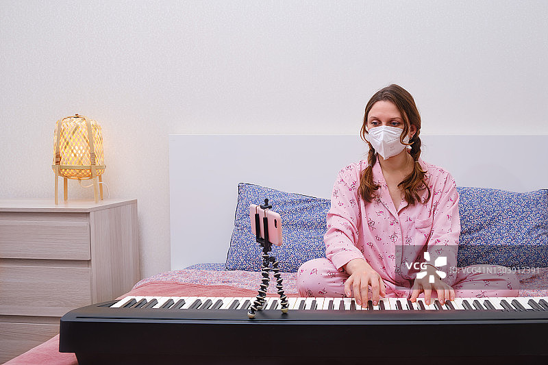 一位戴着医用口罩的妇女坐在家里卧室的床上弹奏钢琴，旁边是电话。隔离音乐课，同时隔离新冠病毒图片素材
