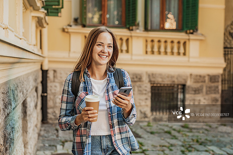 年轻女子使用手机和拿着可重复使用的咖啡杯在街上图片素材