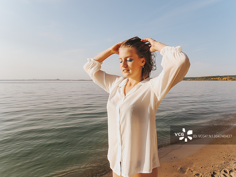 一个快乐的年轻女子穿着白色的衬衫和比基尼在海边日落，和平和宁静图片素材