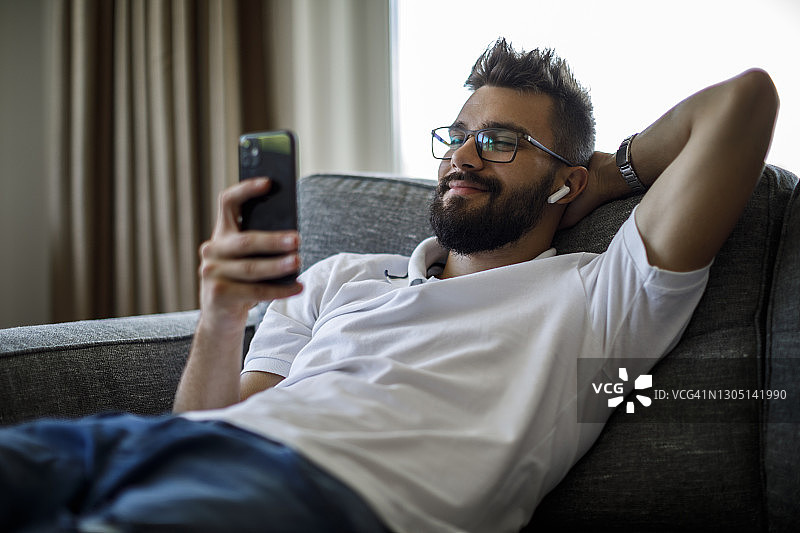 微笑的年轻人用蓝牙耳机在沙发上使用智能手机图片素材