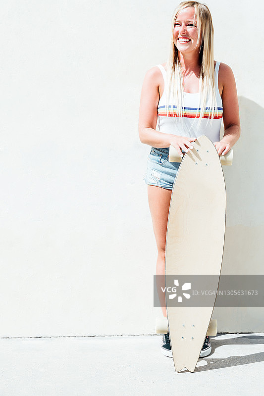 微笑的金发女人在短裤靠在白色的墙壁与她的长板滑板休息在它。图片素材