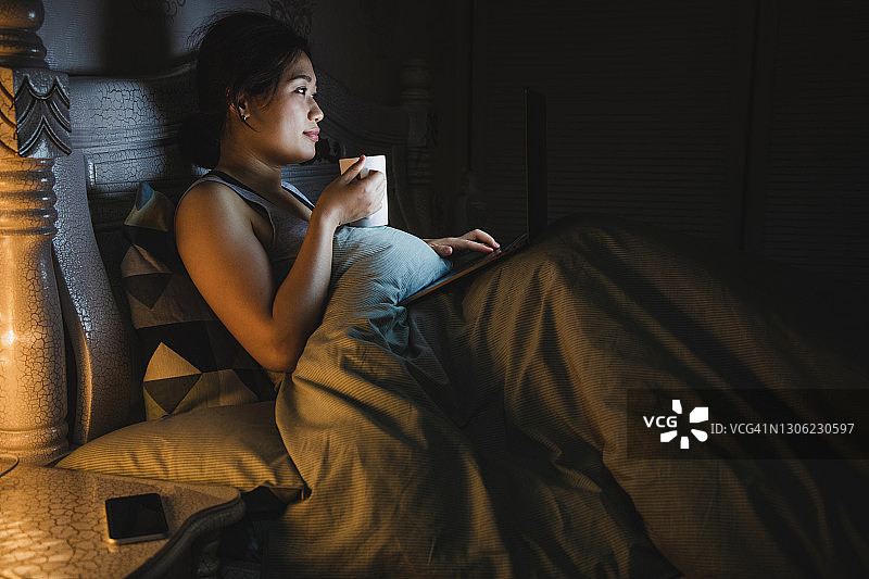 亚洲妇女躺在床上使用笔记本电脑，同时在晚上喝水图片素材