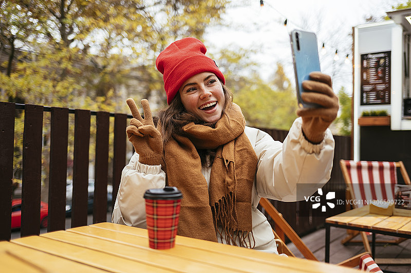 坐在路边咖啡馆用手机自拍，打着和平手势的快乐女人图片素材