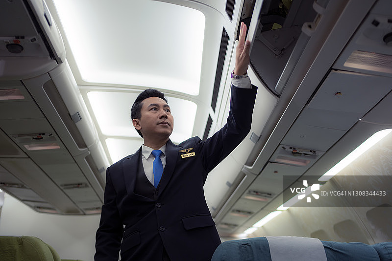 自信的穿着制服的亚洲男性飞行员站在飞机内的乘客座位上。图片素材