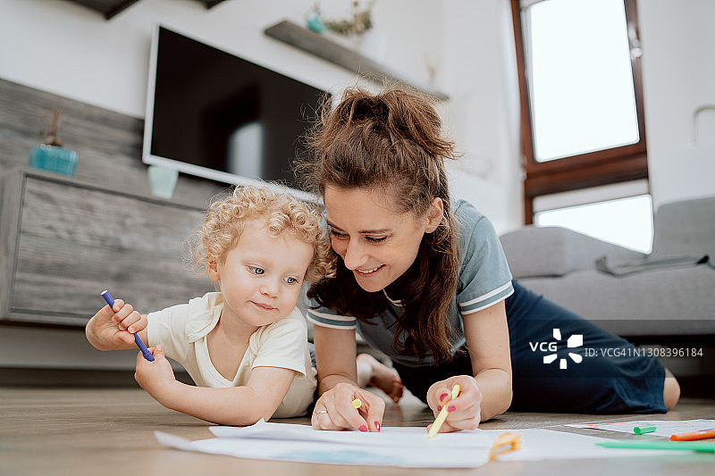 微笑的母亲和她的学龄前的女儿在空闲时间一起画画，而躺在家里客厅的木地板上。妈妈和女儿用马克笔画画图片素材