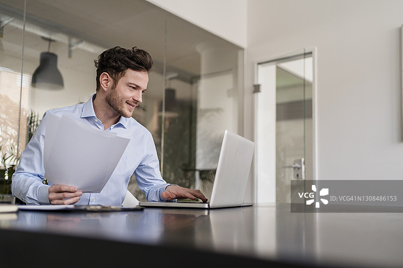 微笑的男性企业家在办公室用笔记本电脑处理文件图片素材