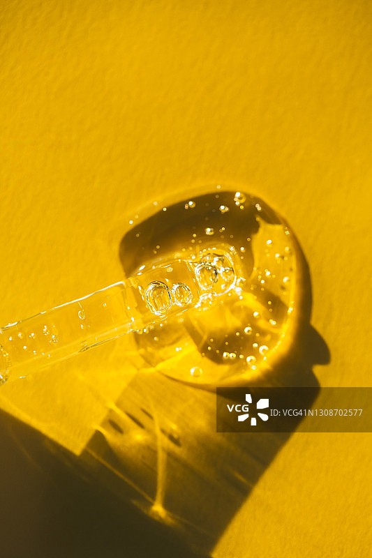 一滴油从玻璃移液管中流出，洒在明亮的黄色背景上。光影的发挥创造了一个抽象的图案。有机身体护理化妆品的概念。2021年的流行色。平躺风格图片素材