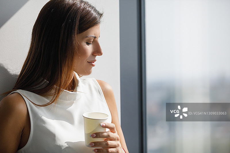 年轻女子在喝咖啡休息时间享受透过办公室窗户的阳光图片素材