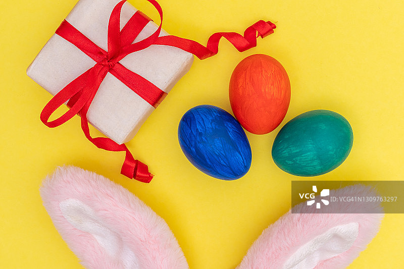 复活节模型:彩色彩蛋，白色礼盒与红色丝带，粉红色兔子耳朵在明亮的黄色背景图片素材