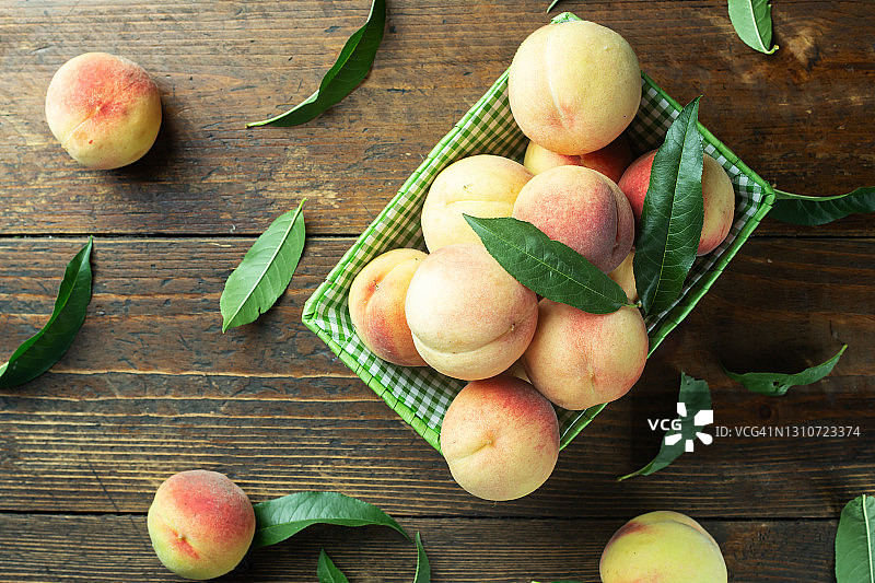 木制背景上的篮子里装着成熟的桃子。桃叶散落在桌面上。图片素材