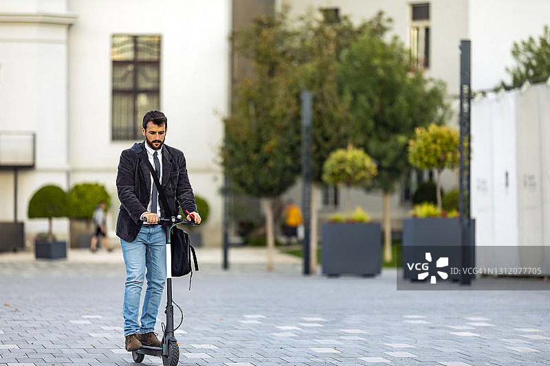 一个自信的年轻商人正骑着滑板车在城市的街道上微笑着。尖峰时刻的年轻商人。图片素材