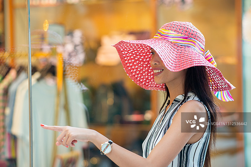 对待自己(购物)。在购物中心的商店橱窗前，一名戴着彩色草帽的亚洲妇女微笑着伸出手指图片素材