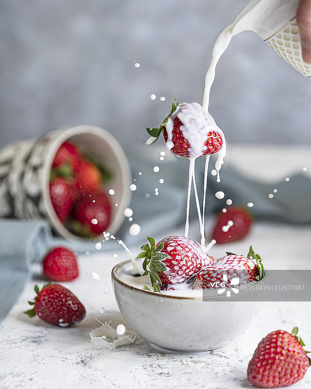草莓掉进牛奶吸管碗里，然后溅到一个有浆果的碗上，桌上是白色的纹理。图片素材