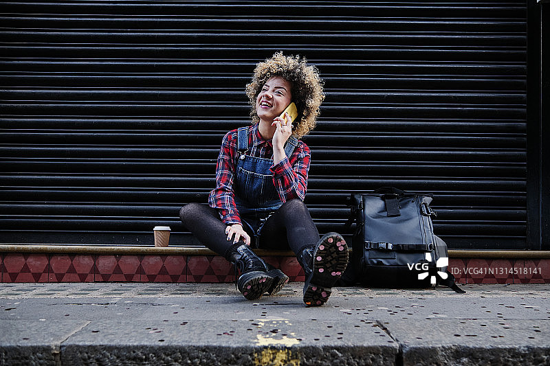 微笑的卷发女人，一边坐在人行道上一边用手机聊天图片素材