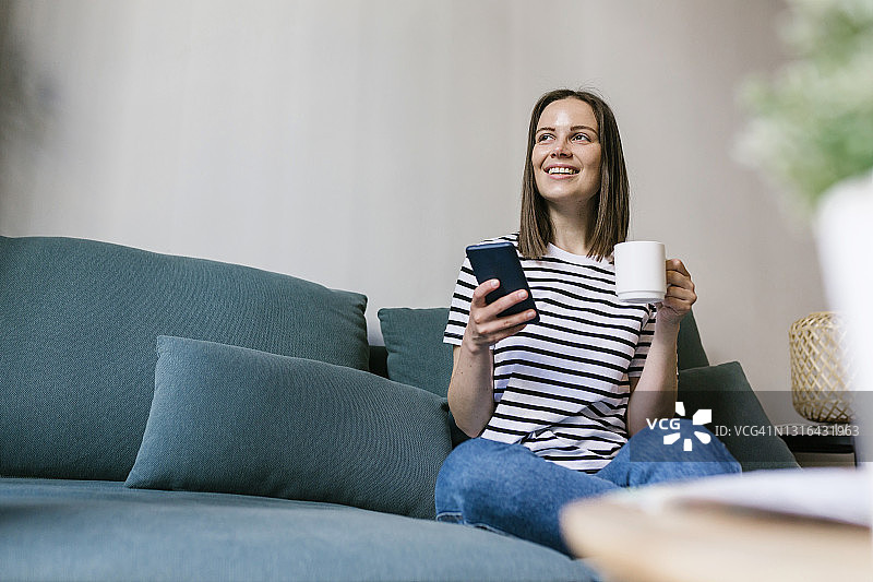 微笑的女人拿着手机在度假屋的沙发上喝咖啡图片素材
