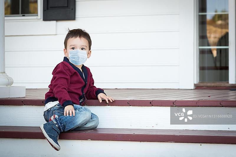 美国冠状病毒大流行期间，一名忧郁孤独的混血男孩戴着医用口罩站在门廊前图片素材