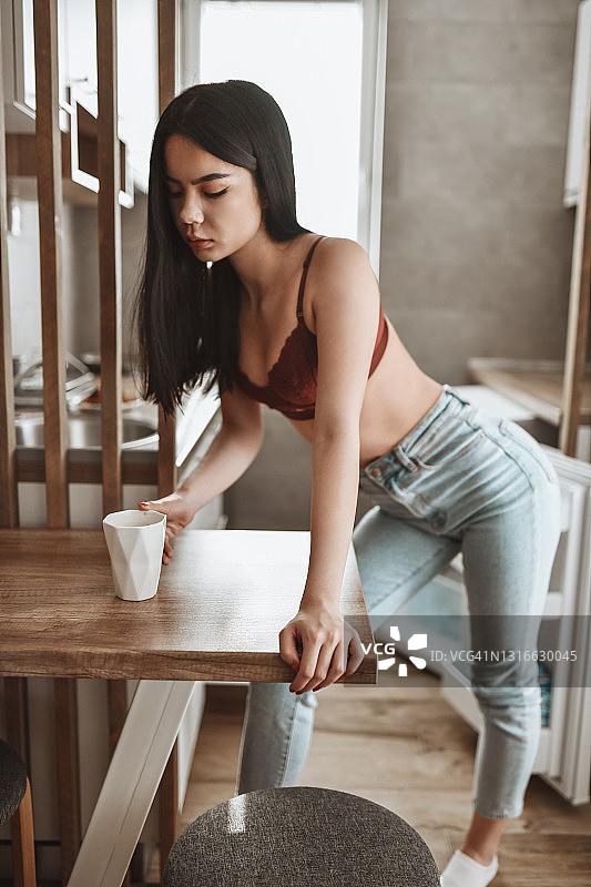 美丽的亚洲女性在胸罩和牛仔裤喝早上咖啡在厨房图片素材