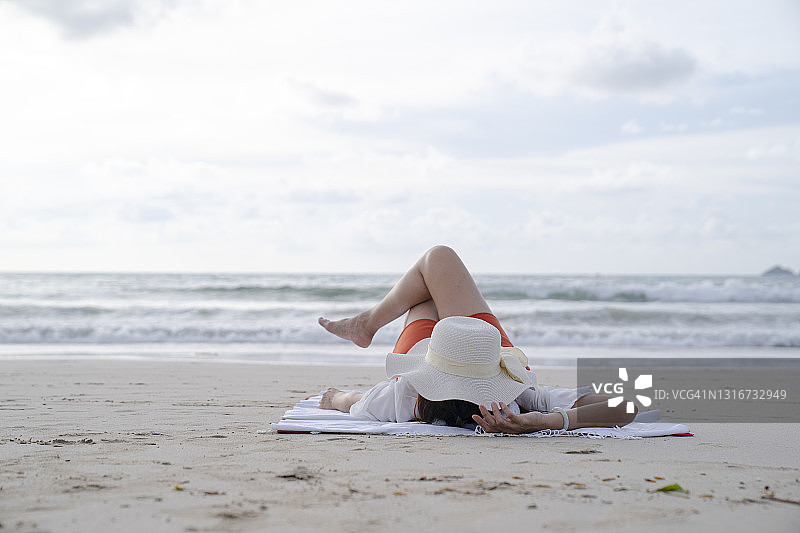 年轻女子伸开双臂躺在垫子上，在海滩上放松和晒日光浴。图片素材