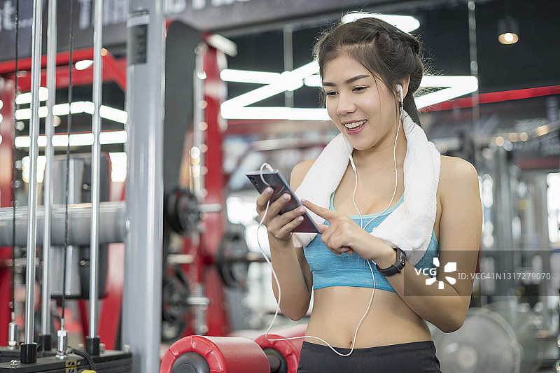 微笑的年轻女子在健身俱乐部用耳机和智能手机听音乐。图片素材