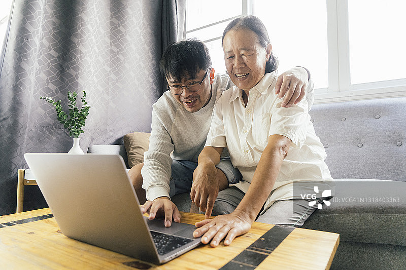 亚洲年轻的成年人儿子教年长的母亲如何使用社交媒体的笔记本电脑在家里图片素材