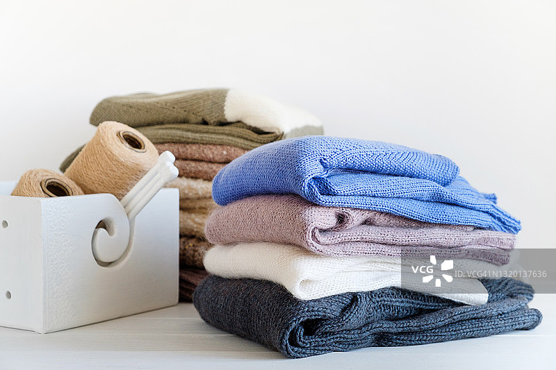 不同颜色的针织品摞在一起，躺在白色的背景上。冬天和秋天暖和的毛衣。在一束束毛线旁边，是编织针。对手工制品的爱好、储存、护理和洗涤的概念。图片素材