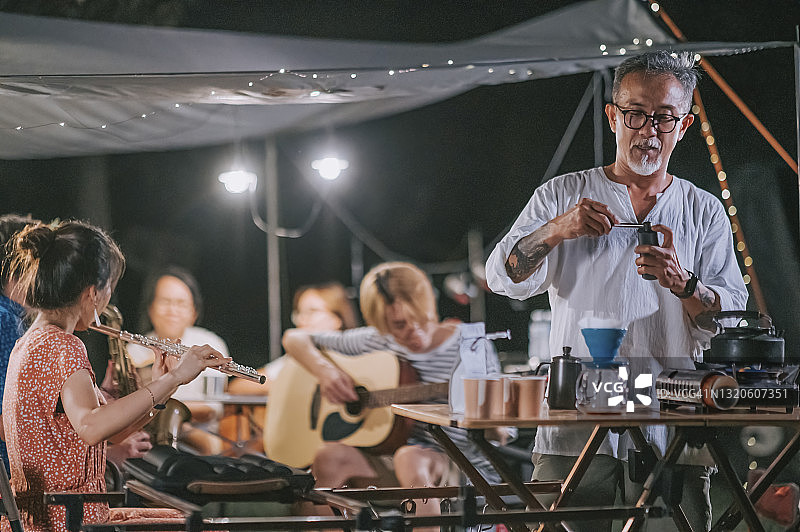 亚洲华人长者磨咖啡，而家人和朋友享受演奏乐器，花园聚会聚会露营帐篷晚上图片素材