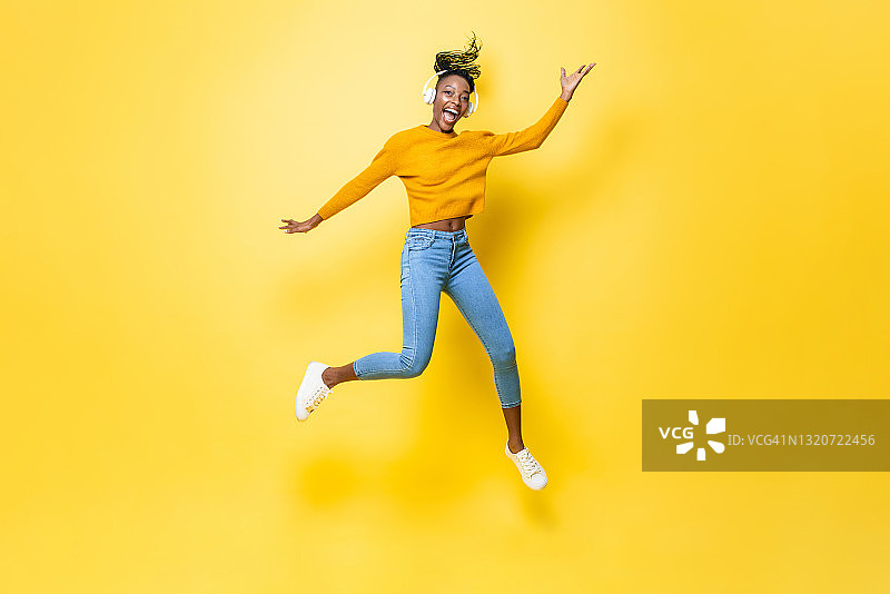 快乐精力充沛的年轻非洲裔美国妇女戴着耳机听音乐和跳跃与手在黄色孤立的演播室背景图片素材