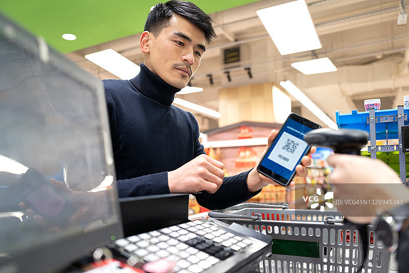 一名亚洲男子在杂货店收银员前通过扫描二维码付款图片素材
