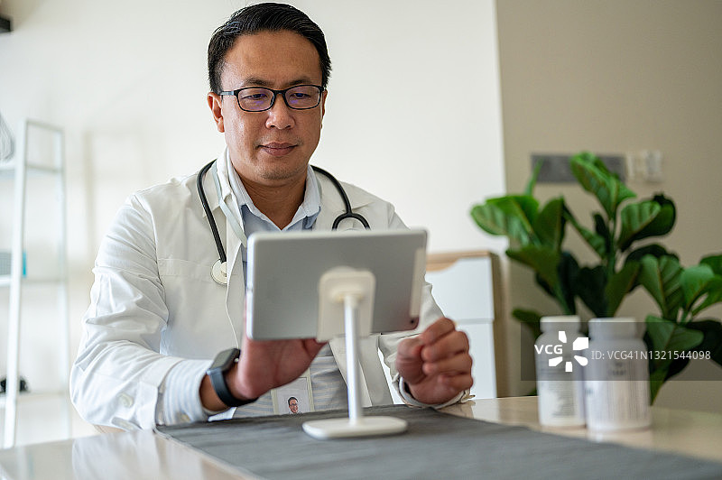 一位亚洲医生坐在诊所里，用笔记本电脑与病人进行在线视频咨询。图片素材
