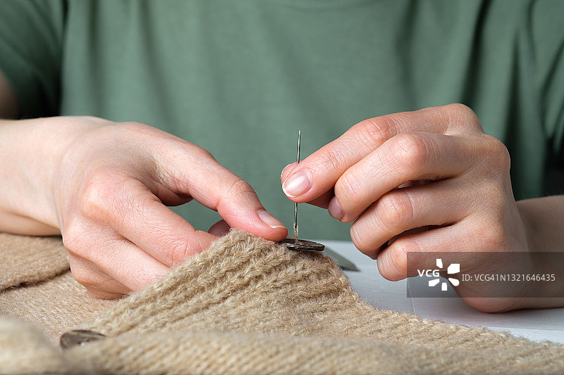 妇女在针织毛衣、开襟羊毛衫或夹克衫上缝一颗钮扣。特写:裁缝、家庭主妇或裁缝用针穿线。一个真实的生活方式的概念，爱好，手工。一个女人和一个母亲的家务责任。图片素材